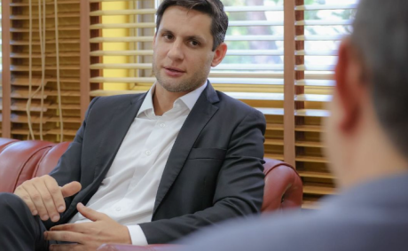 Rafael Motta é branco e tem cabelo castanho. Veste camiseta branca e blazer preto.