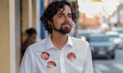 Giovani veste camiseta branca. É branco, tem cabelo cacheado escuro e barba.