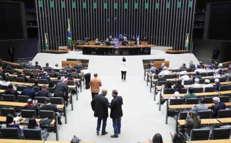 Imagem mostra o plenário da Câmara. é uma sala ampla, chão branco, há muitos assentos dispostos na esquerda e na direita, ocupados por parlamentares. Alguns estão em pé. No fundo, ao centro, há uma mesa de madeira, onde estão dispostos os parlamentares responsáveis por presidir a sessão. Há uma bandeira do Brasil no lado esquerdo.