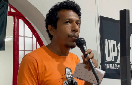 Santiago Belizário (UP) veste uma camisa laranja e segura um microfone. É negro e tem cabelo e bigode pretos.