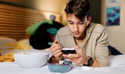 Adolescente deitado na cama mexendo no celular enquanto come salgadinho e doces