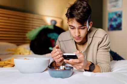Adolescente deitado na cama mexendo no celular enquanto come salgadinho e doces