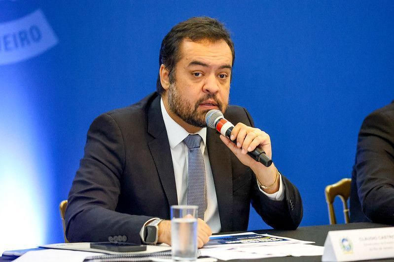 Castro veste paletó preto, camiseta branca, gravata quadriculada e segura um microfone. Está sentado.