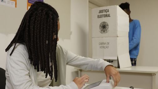 Uma mulher de tranças e roupas claras está sentada em uma mesa e coloca um dedo em uma máquina
