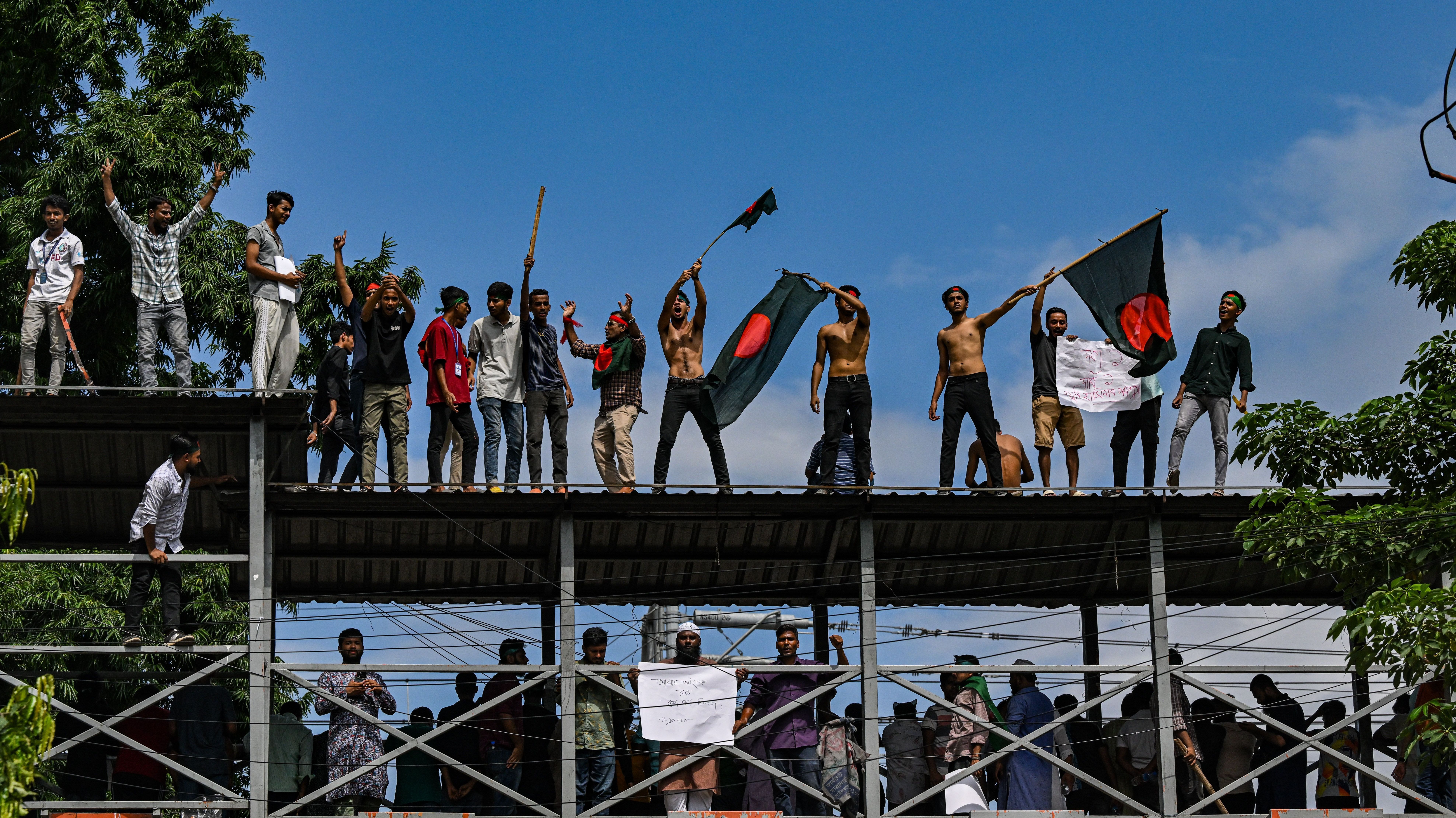 Manifestantes agitam bandeiras nacionais enquanto estão de pé sobre a ponte pedonal em Shahbagh durante um protesto em Dhaka, Bangladesh, em 4 de agosto de 2024, para exigir justiça para as vítimas presas e mortas na recente violência nacional durante os protestos anticotas