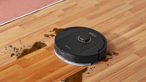 A fotografia mostra um chão sujo e, ao lado, um robô aspirador prestes a limpar o local.