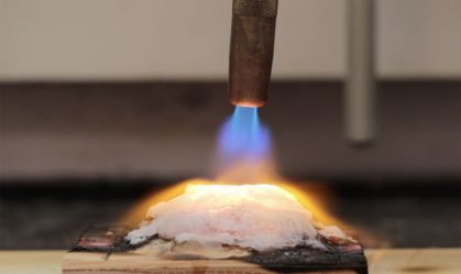 A fotografia apresenta um maçarico colocando fogo em uma madeira compensada envolvida no gel capaz de evitar que casas peguem fogo.