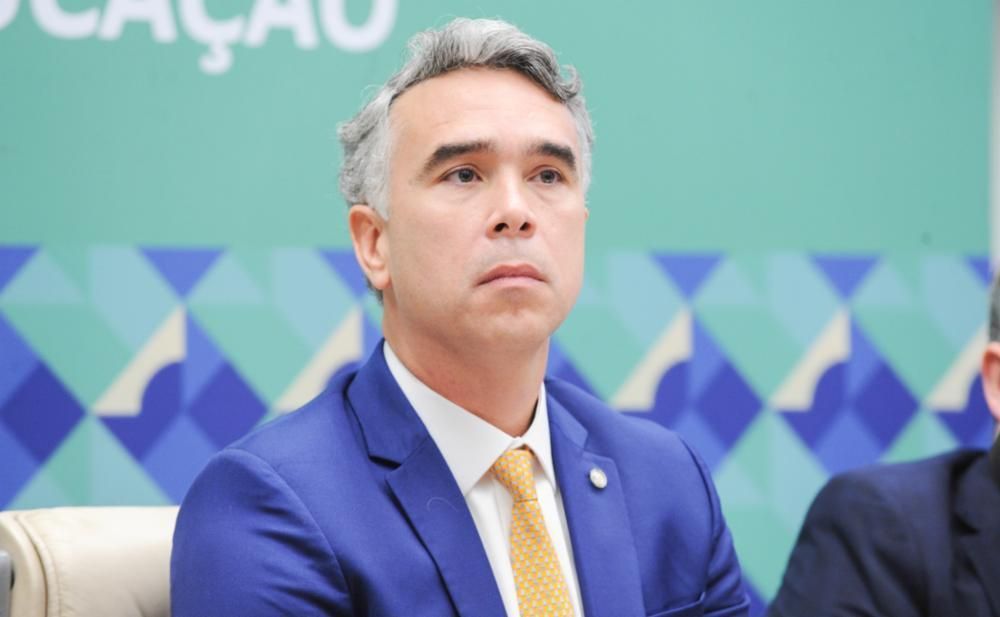 Brito veste terno azul camiseta branca e grava amarela estampada. É branco e tem cabelo grisalho.