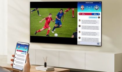 A imagem apresenta uma fotografia de uma TV samsung em um suporte na parede, onde a mnão de um homem aparece segurando um smartphone.