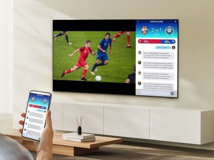 A imagem apresenta uma fotografia de uma TV samsung em um suporte na parede, onde a mnão de um homem aparece segurando um smartphone.