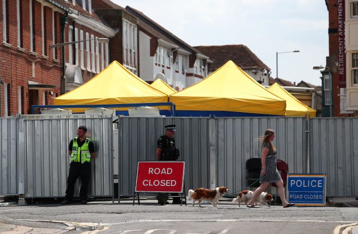 Mulher passa com cachorros por policiais perto de barreiras que bloqueiam a rua de Dawn Sturgess, morta após exposição a um agente nervoso Novichok, em Salisbury, Reino Unido • 19/07/2018 REUTERS/Hannah McKay