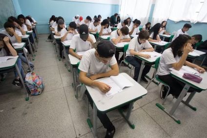 Adolescentes dentro de sala de aula. Eles estão sentados, fazendo uma prova. Todos vestem uma camiseta branca.