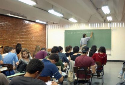 Sala de aula. Há alunos no local. O professor está escrevendo na lousa.
