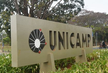 Placa em que está escrito "Unicamp". Ela está no centro da imagem e é cinza.