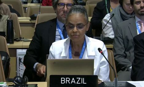 Imagem mostra a ministra do Meio Ambiente e Mudanças Climáticas Marina Silva, discursando no final da COP 29. Ela usa um blazer branco, óculos e tem o cabelo preso em um coque. Uma placa com Brasil escrito com Z está posicionada a frente dela.