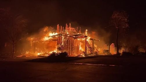 Imagem mostra estrutura de casa destruída em meio a chamas de noite.