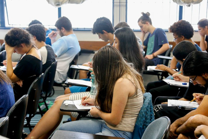 Estudantes dentro da sala de aula. Eles estão sentados e fazem uma prova.