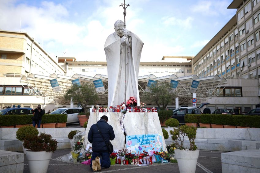 Homem reza perto de estátua do papa João Paulo 2º no Hospital Gemelli, em Roma, onde o papa Francisco está internado. • Reuters/Alkis Konstantinidis