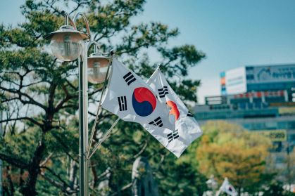 Bandeira da Coreia do Sul. Ela é branca, com um círculo vermelho e azul no centro. Há árvores no fundo.