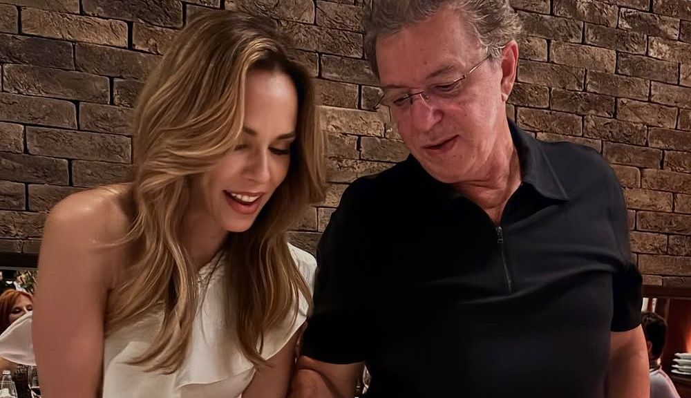 Ana Furtado e Boninho cortando bolo. Ana usa um vestido branco e Boninho uma roupa preta.