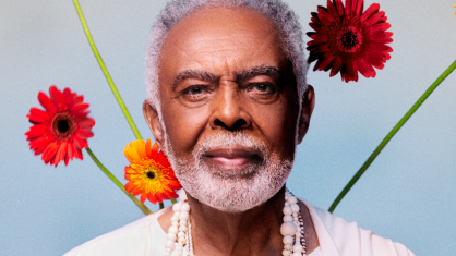 O cantor Gilberto Gil. Ele veste uma camisa branca. Há flores atrás dele.