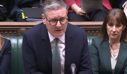 Keir Starmer discursando no Parlamento Britânico. Elé é branco e tem o cabelo grisalho. Usa um terno azul escuro, camisa branca, gravata verde e óculos de grau.