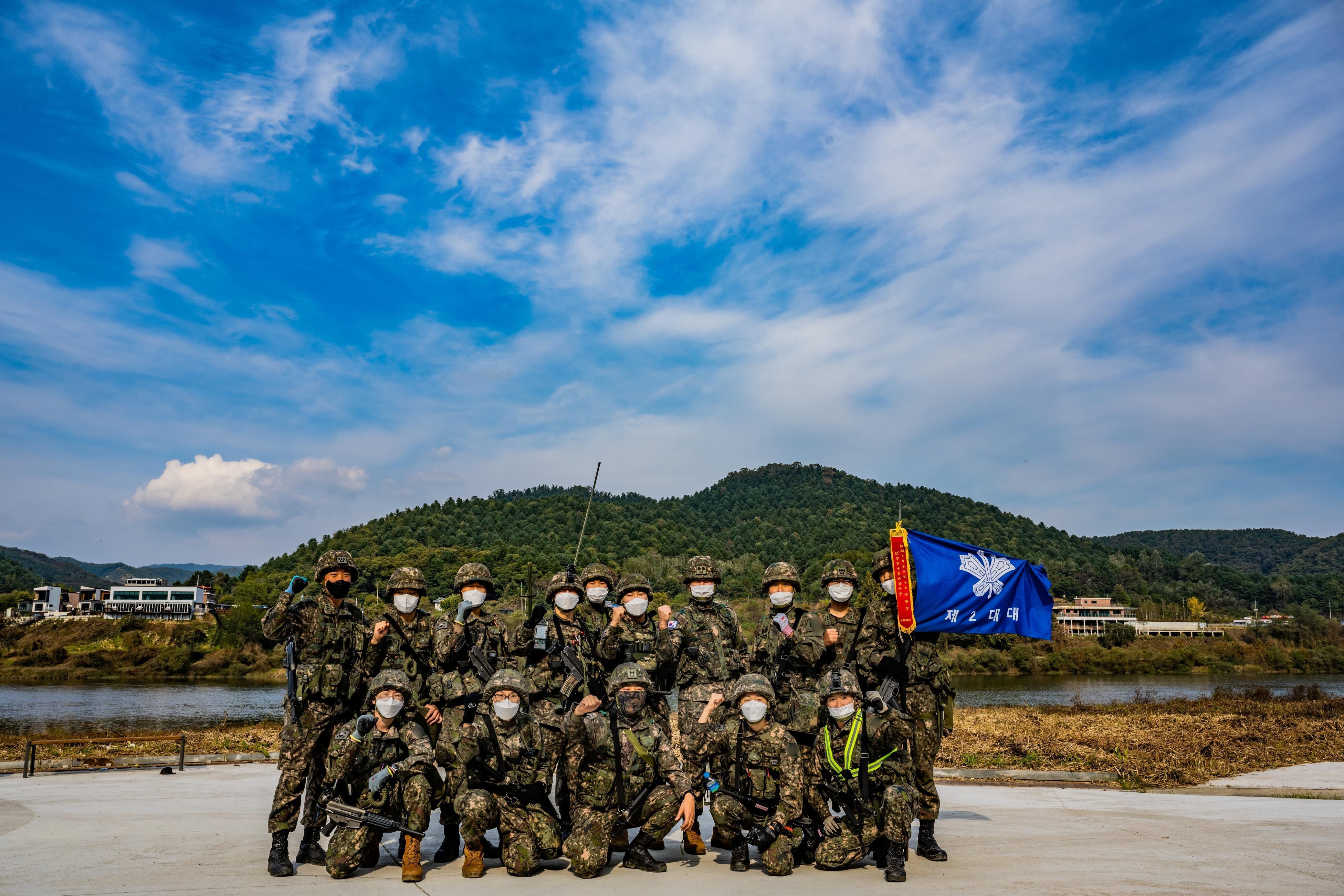 Samuel Jun ao lado de seus companheiros de pelotão, todos em uniforme militar e equipados, posando para a foto ao ar livre. • Arquivo Pessoal