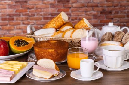 Mesa de café da manhã repleta de alimentos variados, incluindo pães, doces, bolo, iogurte, chá, café, suco de laranja, presunto, queijo, mamão e maçã