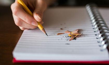 Pessoa escrevendo em um caderno. Há ponta de lápis em cima da folha
