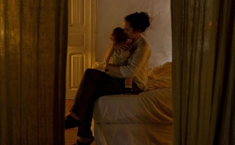 Na imagem, mulher sentada na cama com as pernas cruzadas segura bebe no colo.