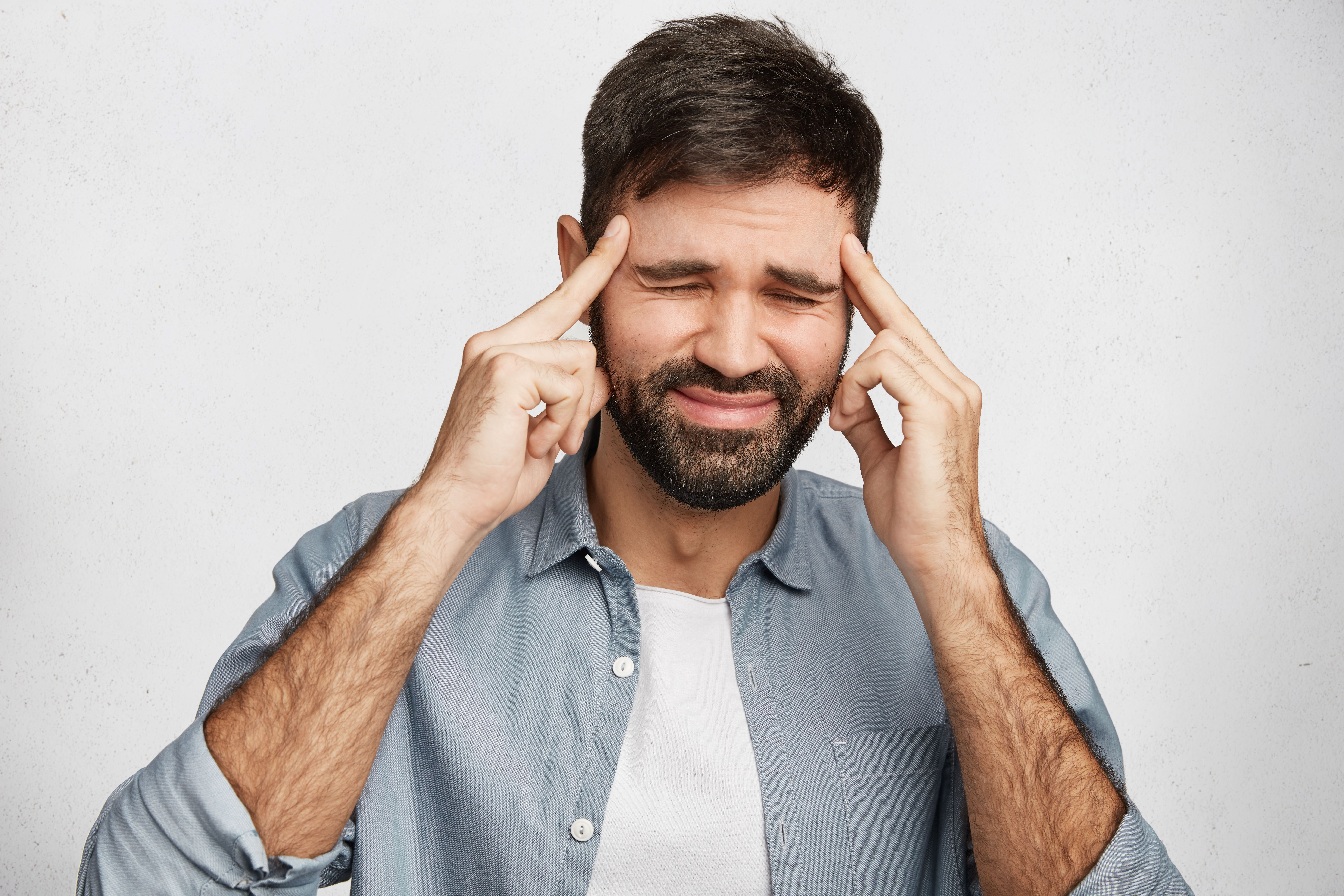 Stressful brunet young bearded male keeps fingers on temples tries to concentrate and find solution in mind, closes eyes, has unhappy expression, isolated over white background. Depression concept