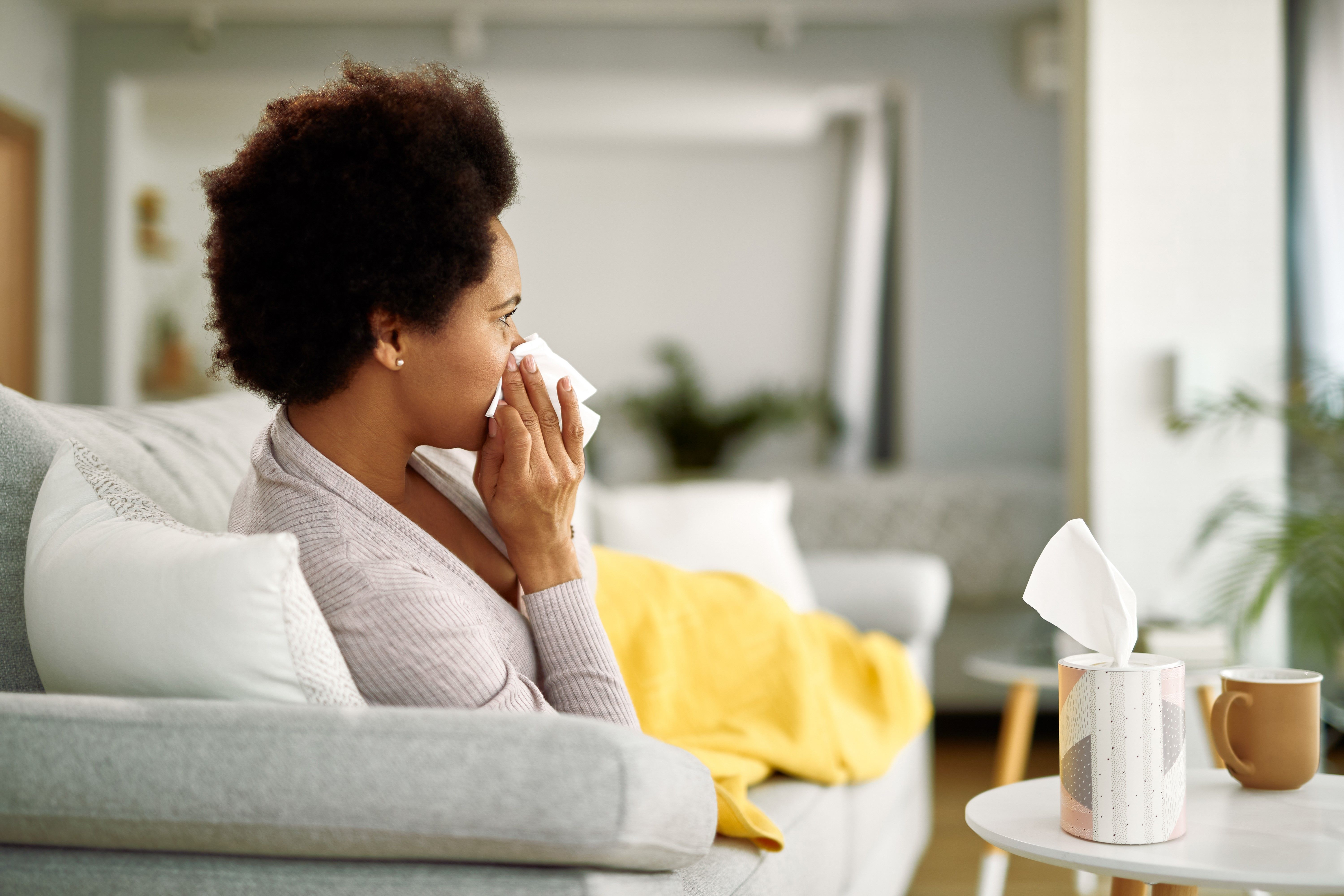 African American woman with flu virus blowing nose while resting on the sofa.