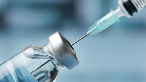 A imagem mostra uma seringa e a enfermeira pegando o produto no recipiente que contém o remédio