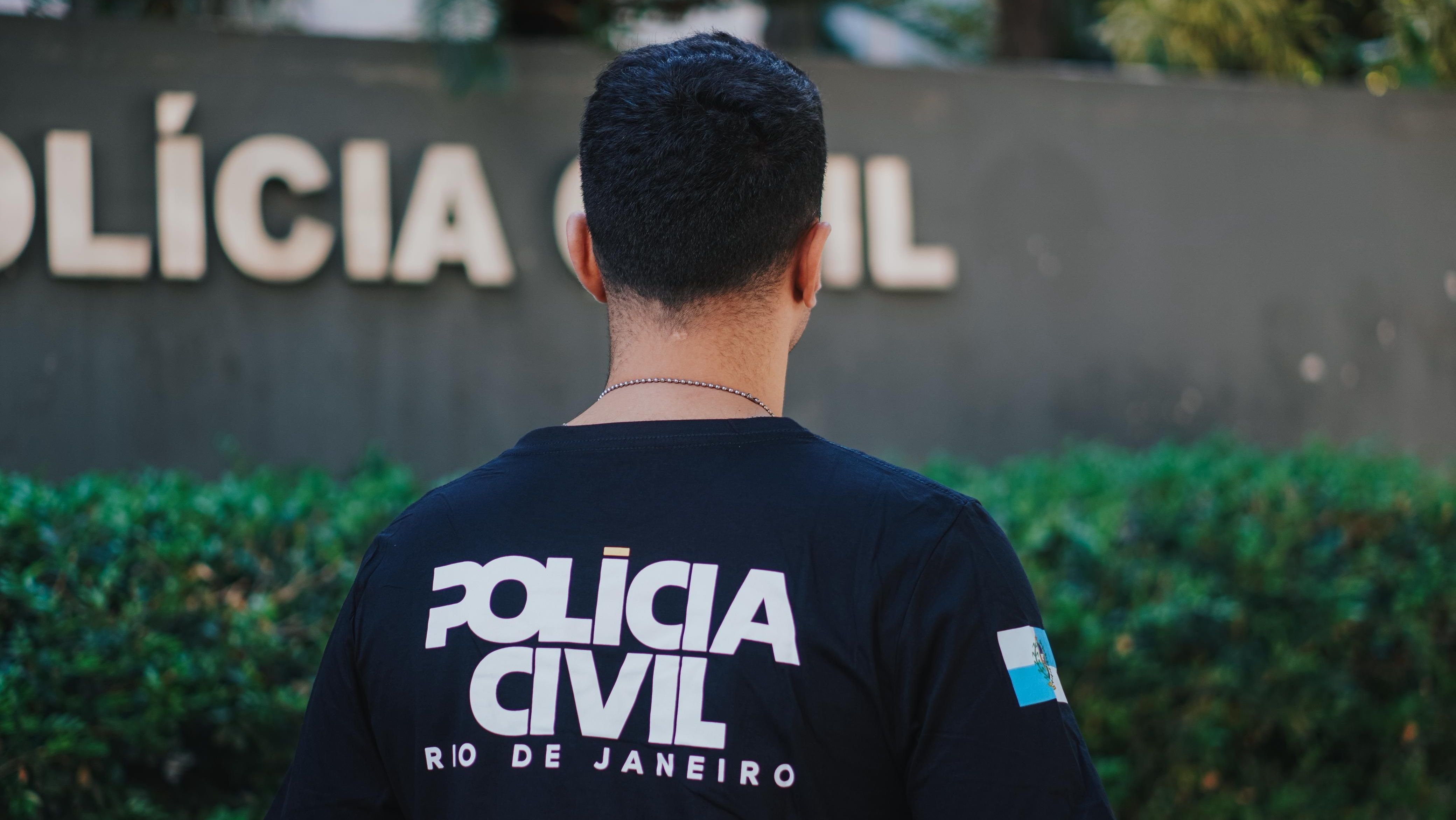 Policial civil de costas com uma blusa preta e um fundo desfocado da delegacia com arbustos