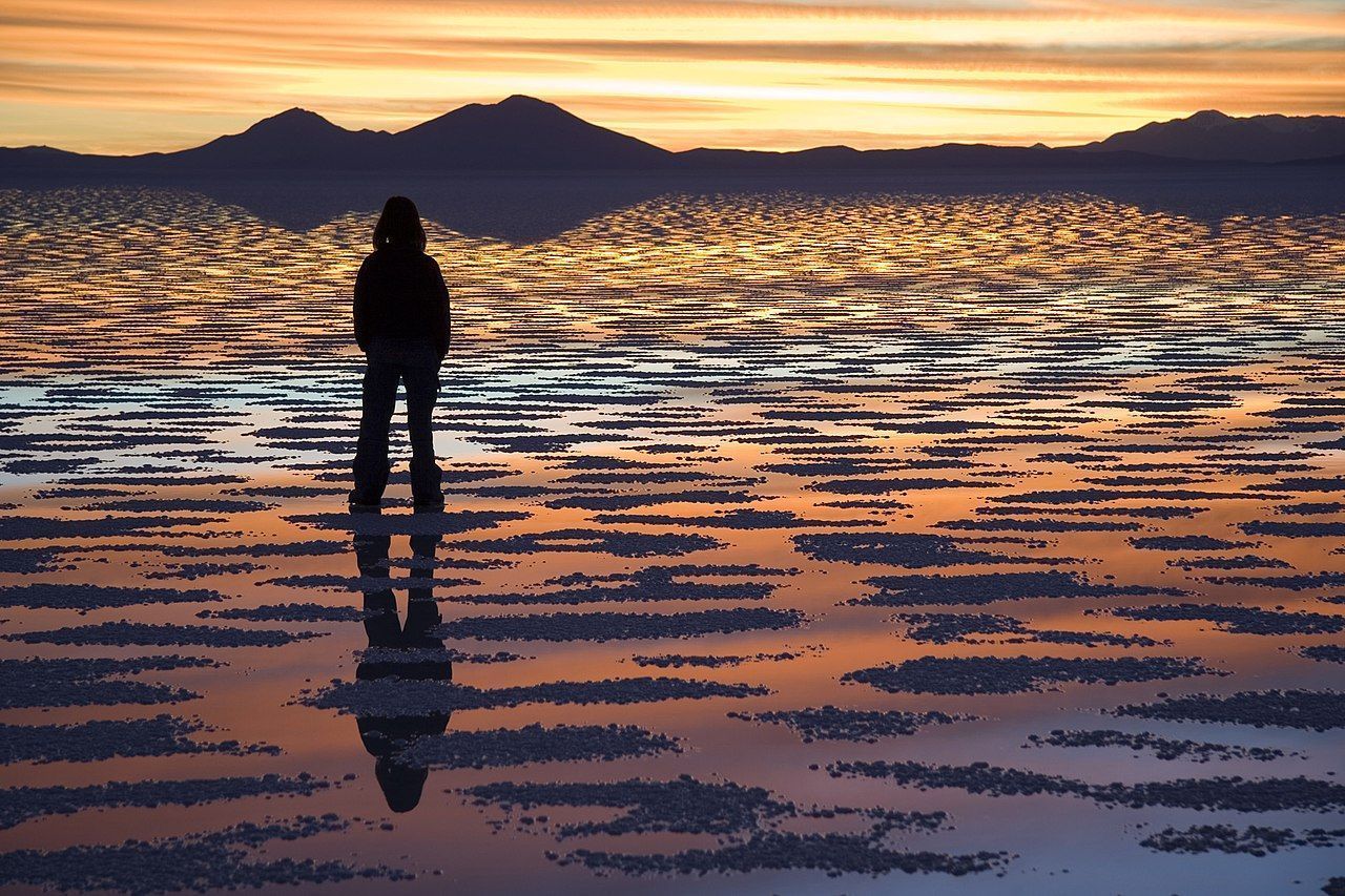 A fotografia mostra uma pessoa em pé no Salar de Uyuni, demonstrando o efeito de espelho natural causado em estações chuvosas.