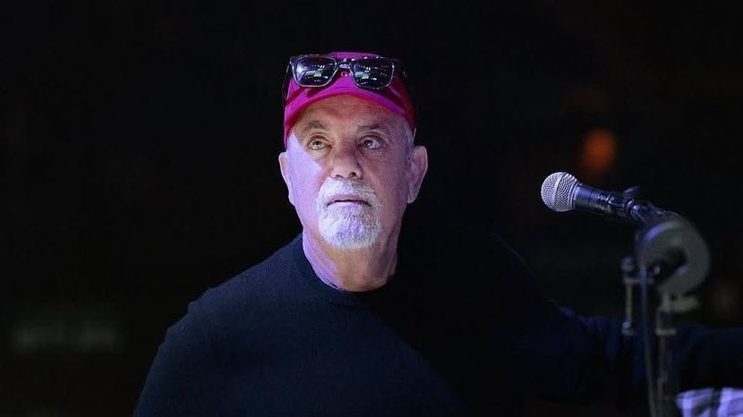 Billy Joel em palco, segurando um microfone, sorridente após cirurgia