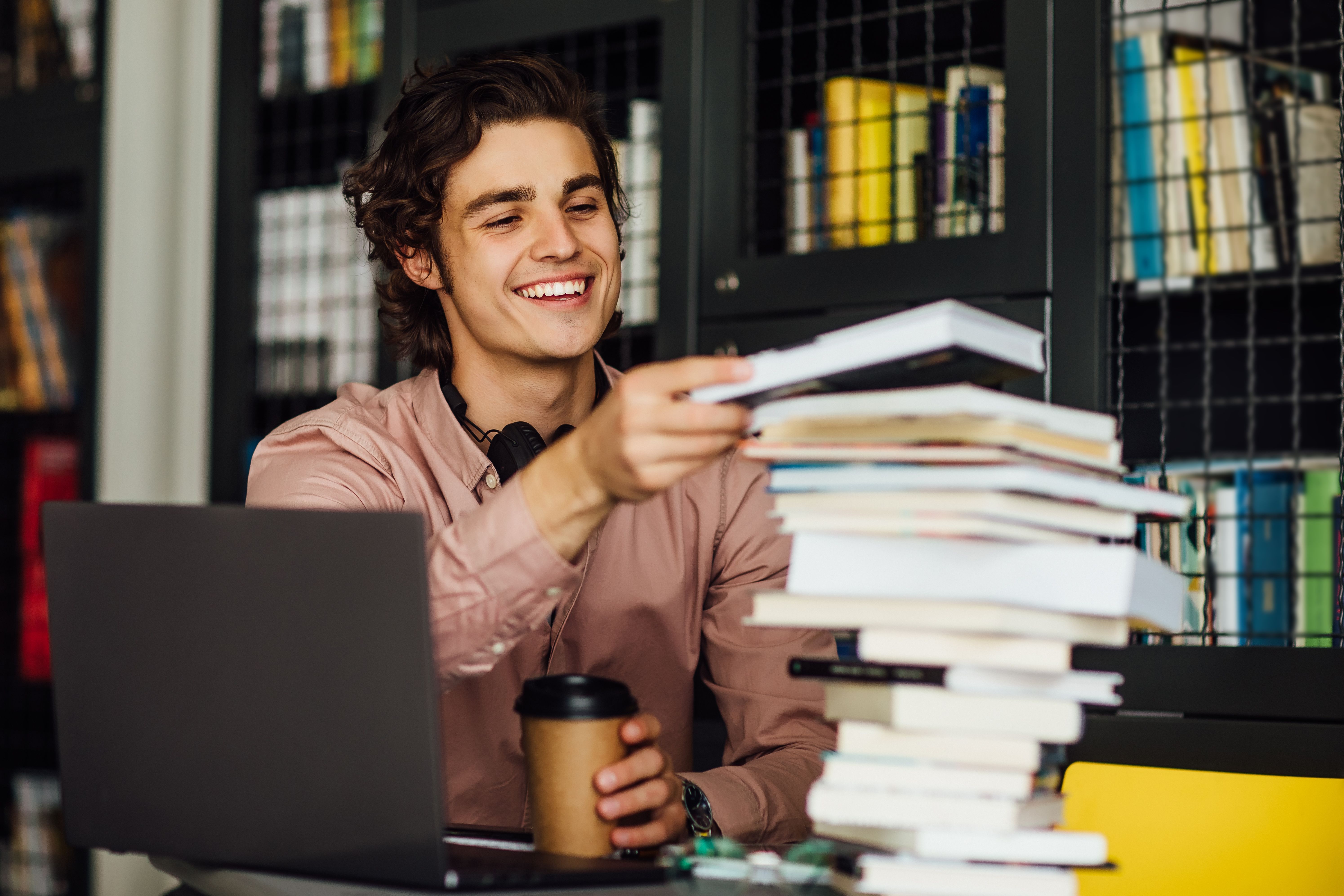 Um jovem pega um livro de uma pilha de livros de sua mesa. Ele segura um copo de café em uma das mãos