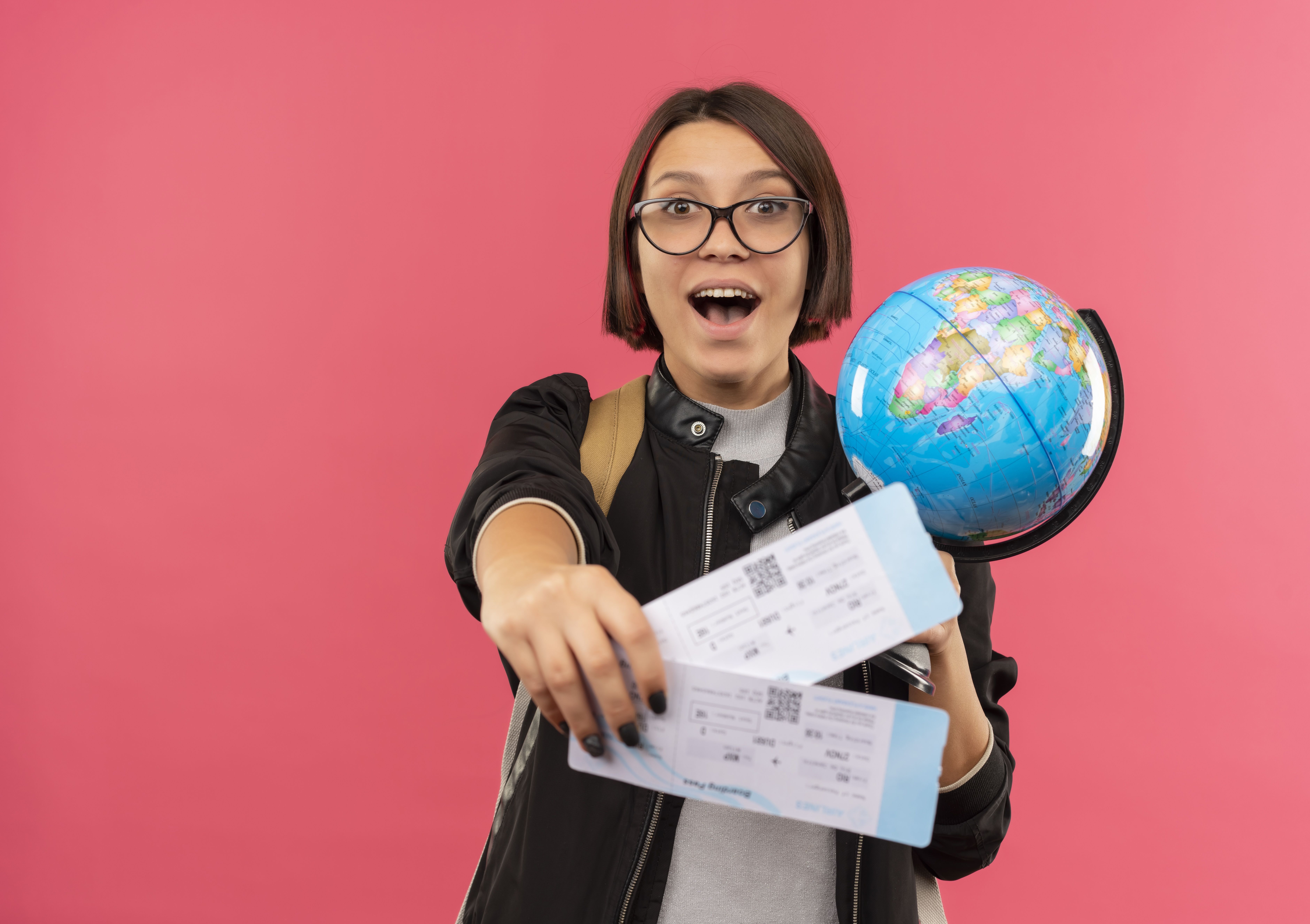 impressed young student girl wearing glasses and back bag stretching out airplane tickets and holding globe isolated on pink background with copy space