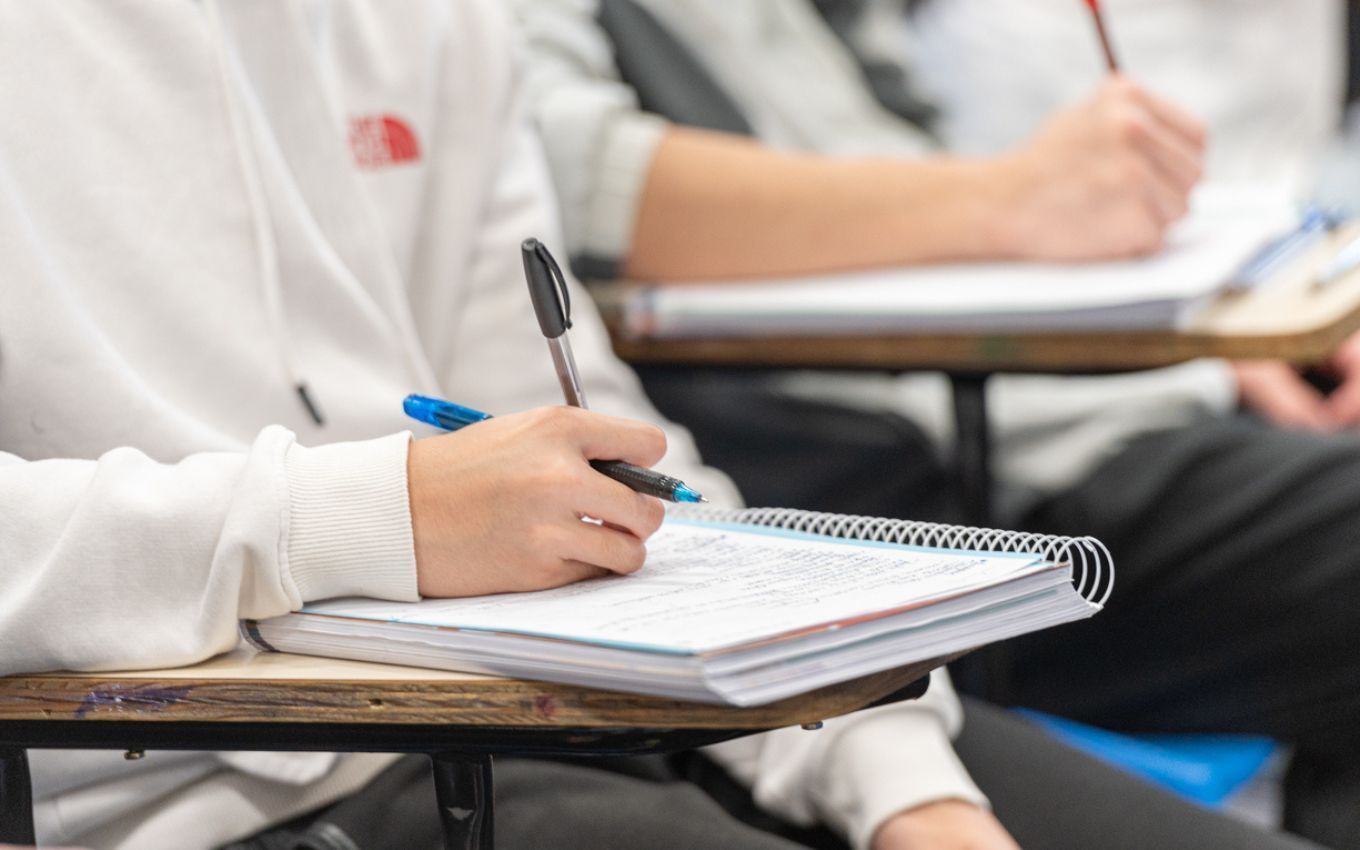 Estudantes sentados em carteiras escolares, em foco um aluno escrevendo em um caderno sem ter seu rosto exposto