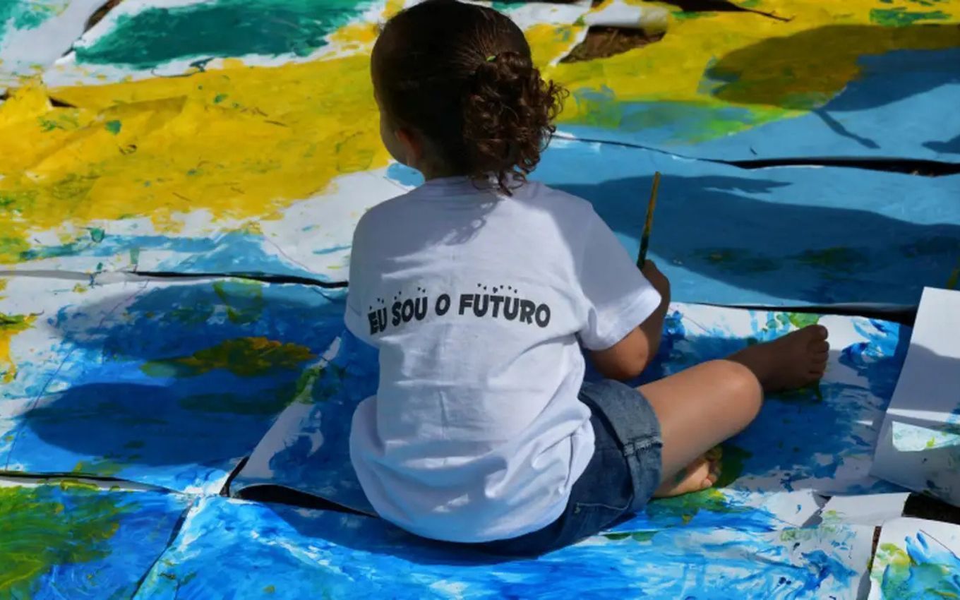 Criança sentada no chão com as cores da bandeira brasileira e com uma camiseta branca escrita Eu Sou o Futuro na cor azul