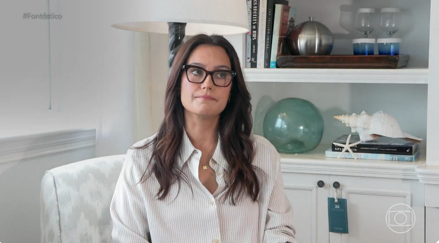 Emma Heming Willis está sentada usando uma camisa branco e óculos escuros em uma sala