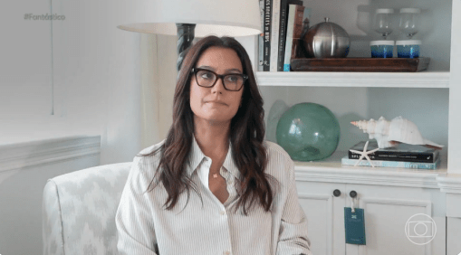 Emma Heming Willis está sentada usando uma camisa branco e óculos escuros em uma sala