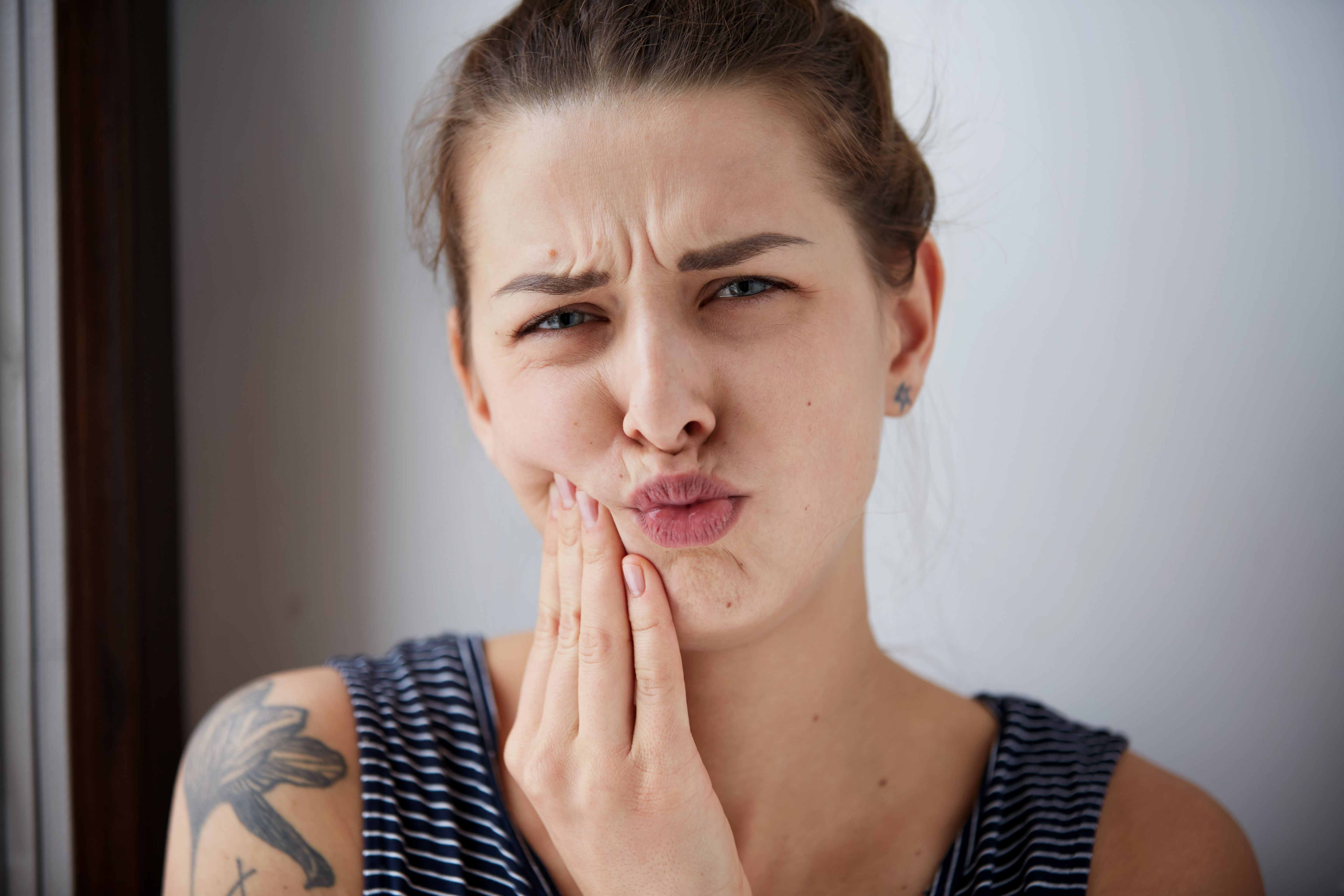Closeup portrait of young woman with sensitive toothache crown problem about to cry from pain touching outside mouth with hand, isolated grey background. Negative emotion facial expression feeling.