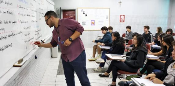 Professor escreve em quadro branvo, enquanto alunos observam