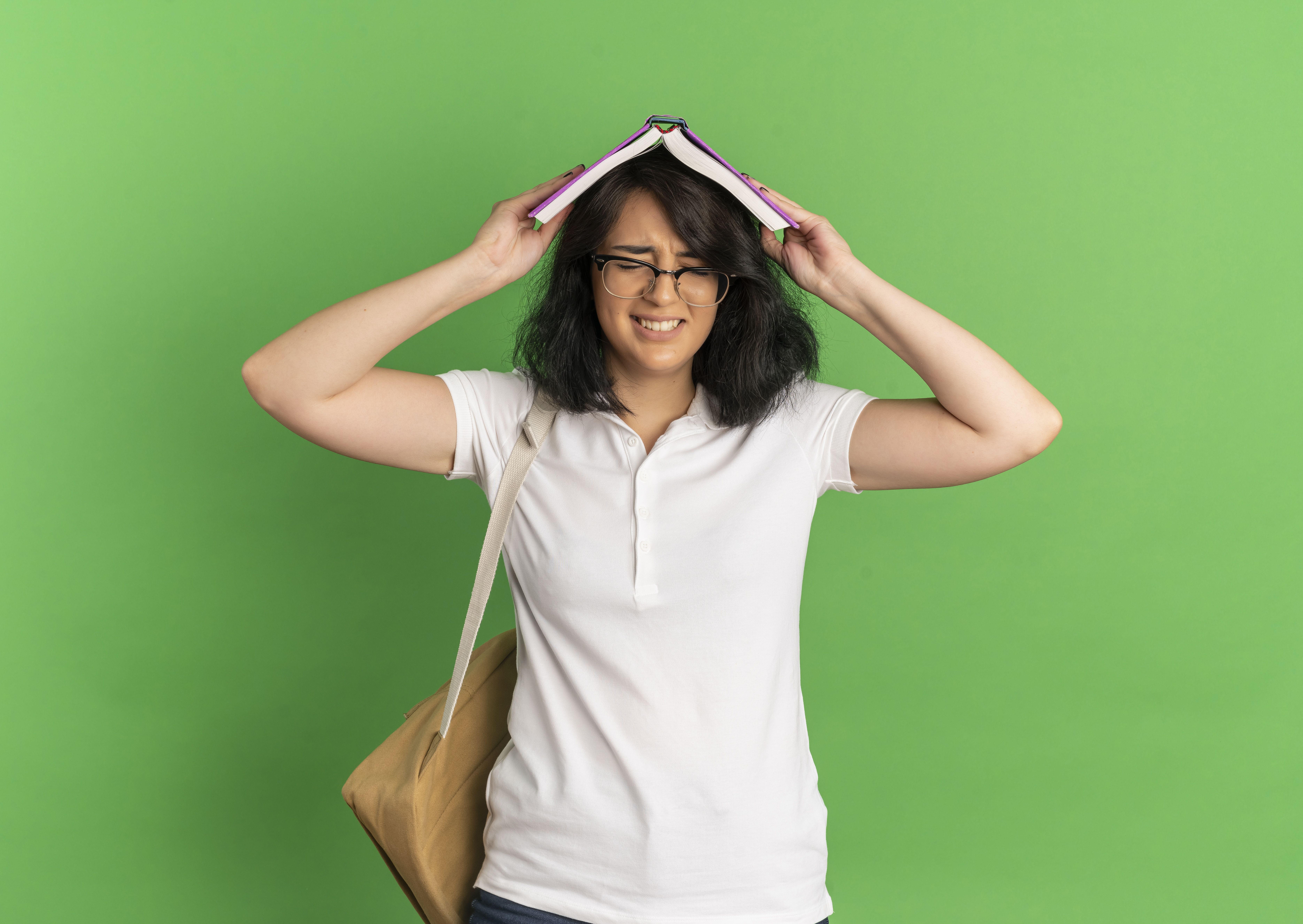 young annoyed pretty caucasian schoolgirl wearing glasses and back bag holds book over head isolated on green background with copy space