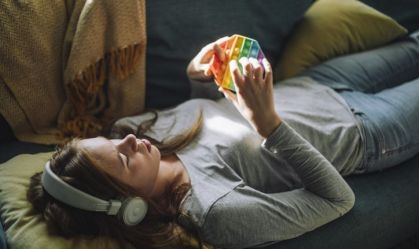 Teenage girl listening to music and playing with push pop toy while lying down on sofa at home