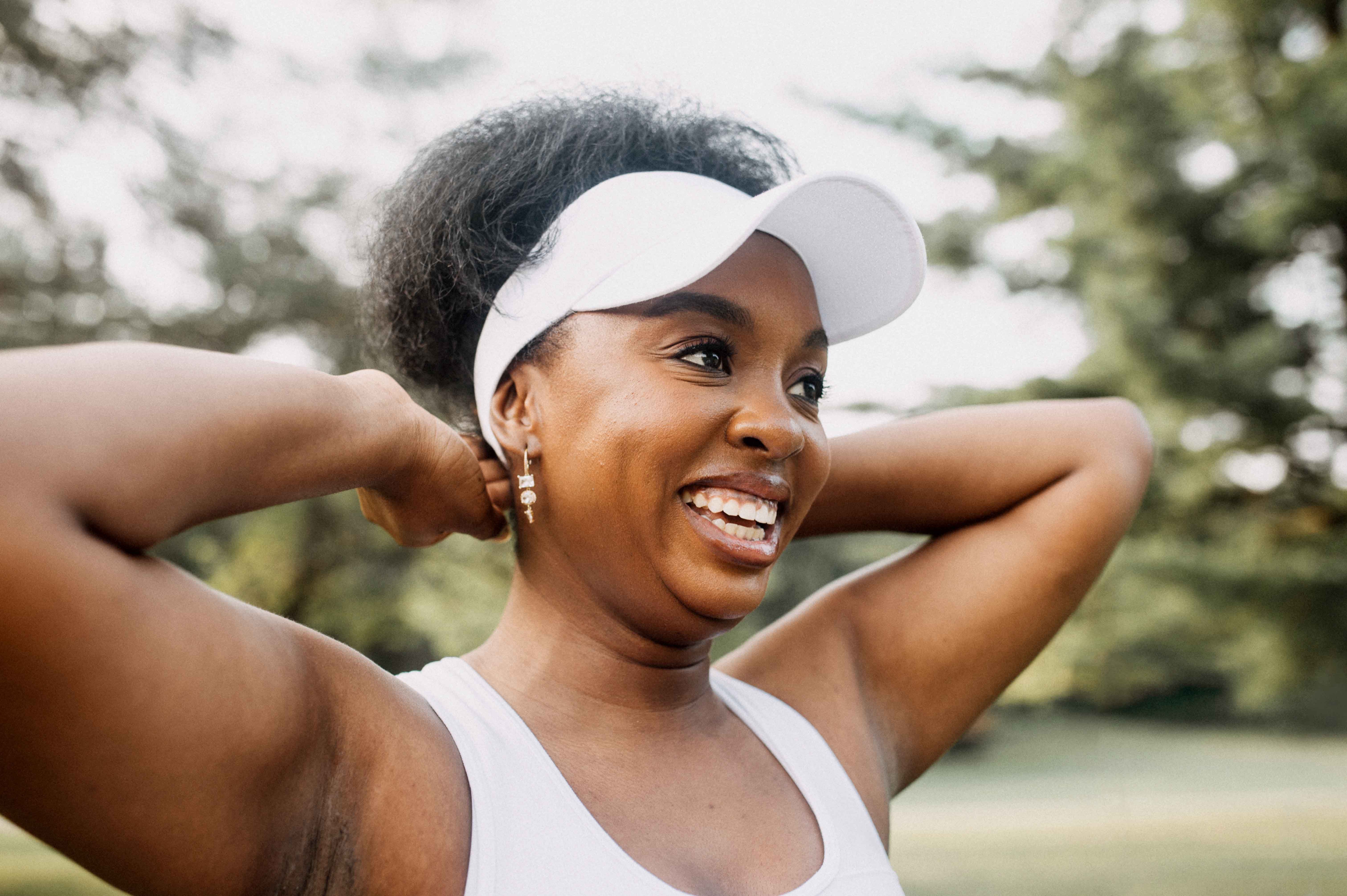 A woman, in athletic attire, is smiling broadly as she stretches her arms behind her head, enjoying an outdoor setting.