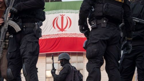 Agentes armados das forças especiais da polícia iraniana monitoram uma área enquanto estão em um veículo militar blindado em frente a uma bandeira do país durante uma manifestação pró-governo no centro de Teerã, Irã, em 12 de janeiro de 2026