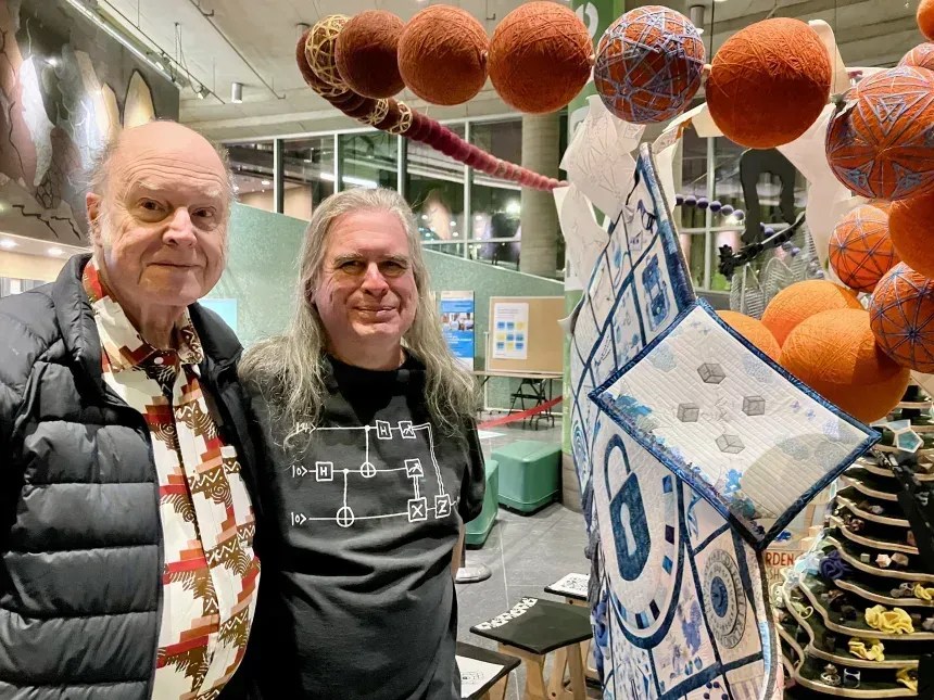 Bennett e Brassard posam em 2024 com a pequena colcha de retalhos de criptografia quântica , que retrata de forma fantasiosa como funciona o protocolo BB84, no Mathemalchemy da Universidade de Quebec em Montreal • Lise Raymond