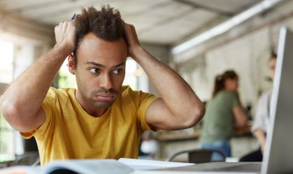 Stressed young African American student of school of economics feeling frustrated while working on diploma project, sitting at coworking space in front of open laptop computer, holding head with hands